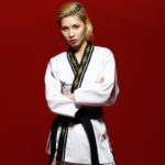Jeune femme blonde, porte un uniforme de taekwondo blanc avec le col doré et une ceinture noire, ses bras sont croisés