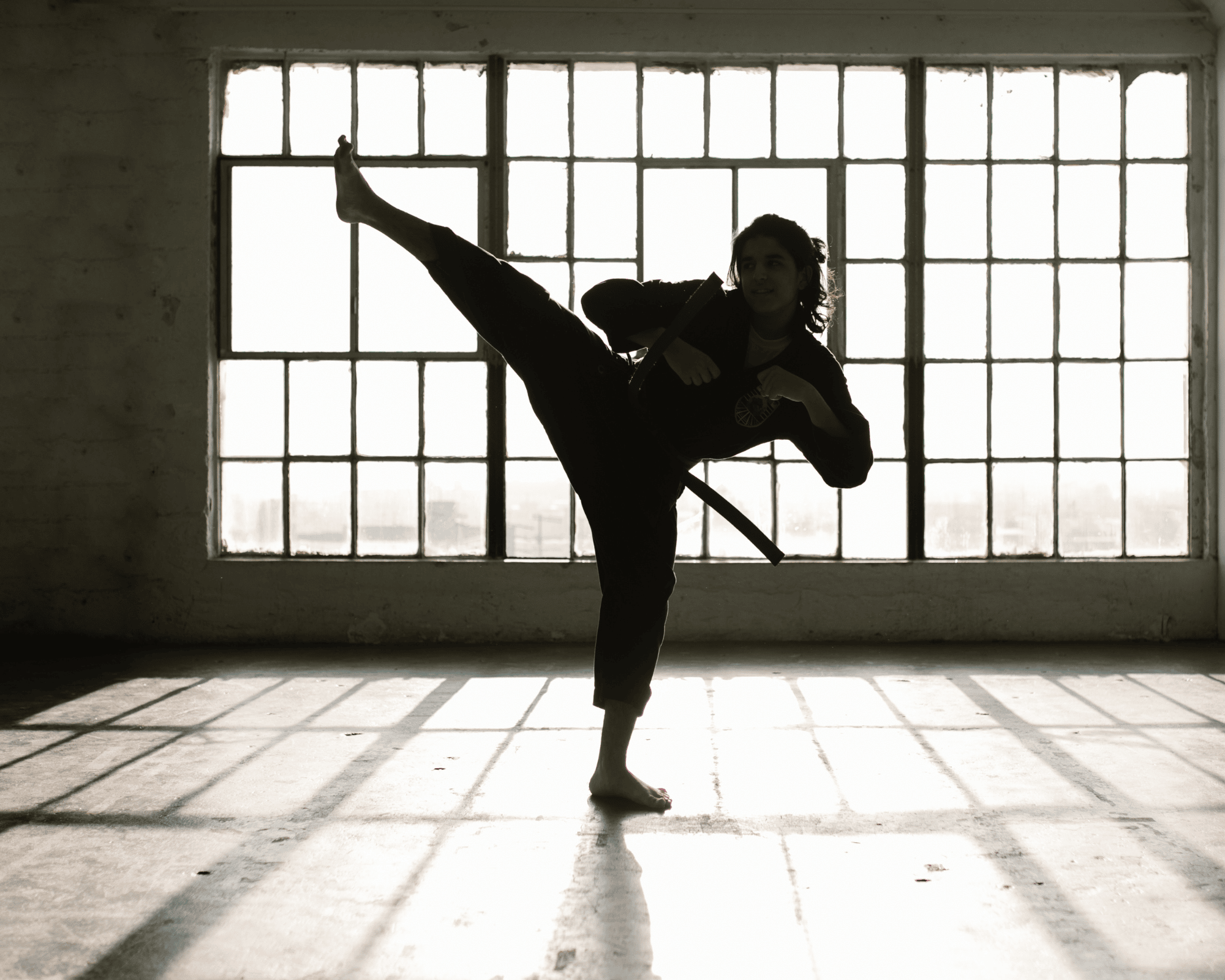 Silhouette de femme en train de lever sa jambe comme dans un combat d'art martial, on la voit dans une salle vide devant des fenêtre du style des salles de sports d'arts martiaux