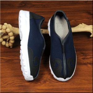 paire de chaussures d'arts martiaux, bleues avec des motifs en simili cuir noir, et semelle blanche, une des chaussures est présentée à plat et l'autre sur le côté, elles sont appuyées sur un morceau de bois