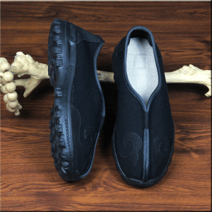 paire de chaussures d'arts martiaux, noires avec des motifs en simili cuir noir, et semelle noire, une des chaussures est présentée à plat et l'autre sur le côté, elles sont appuyées sur un morceau de bois