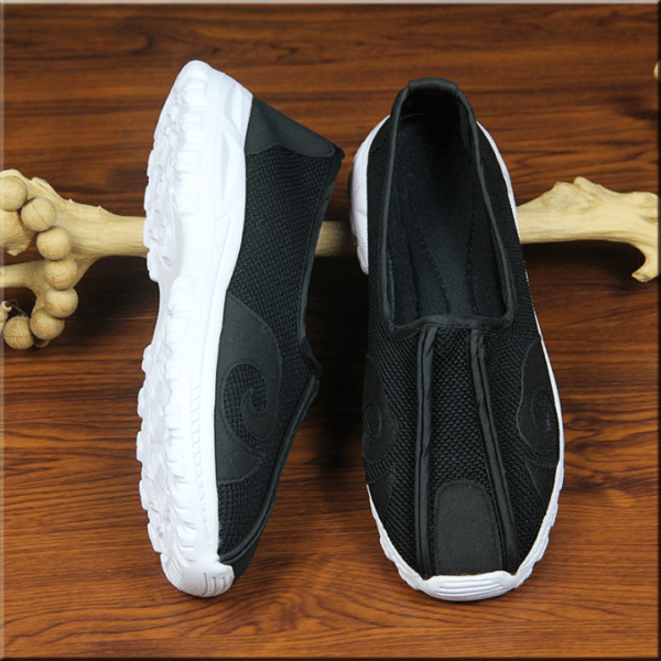 paire de chaussures d'arts martiaux, noires avec des motifs en simili cuir et un double pli sur le devant noir, et semelle blanche, une des chaussures est présentée à plat et l'autre sur le côté, elles sont appuyées sur un morceau de bois