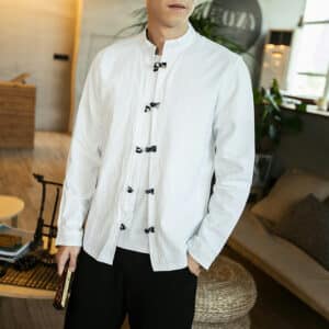 Mannequin homme debout dans une pièce, portant un t-shirt blanc et par-dessus une chemise chinoise ouverte avec fermeture à boutons traditionnels foncés presque noirs. On ne voit pas sa tête, ni ses jambes, il a un éventail fermé dans une main et l'autre dans la poche de son pantalon.