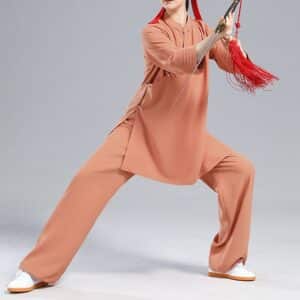 Mannequin femme dans une posture de Tai-chi, portant des baskets blanches avec la semelle orange, un pantalon ample orange et une tunique orange également, fendue sur les côtés, à manches trois-quarts. Elle pose sur un fond gris clair.