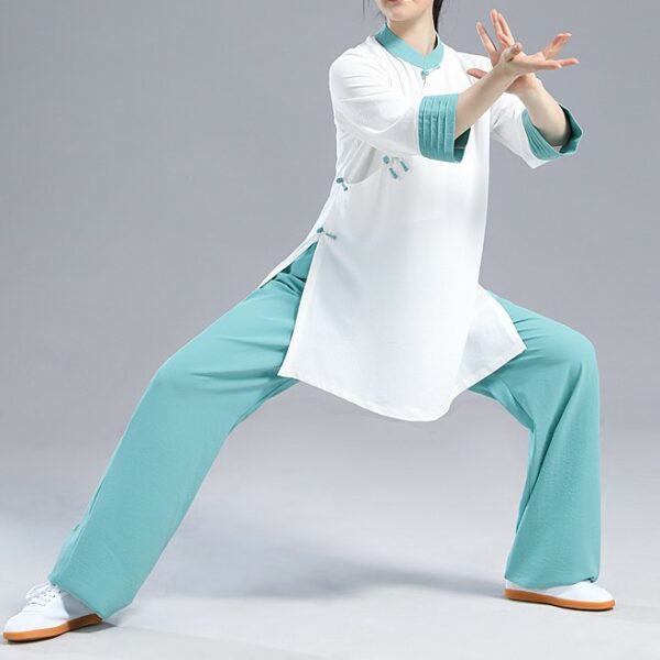 Mannequin femme dans une posture de Tai-chi, portant des baskets blanches avec la semelle orange, un pantalon ample bleu et une tunique blanche avec une finition bande bleue au niveau du col, fendue sur les côtés, à manches trois-quarts finissant par une bande bleue. Elle pose sur un fond gris clair.