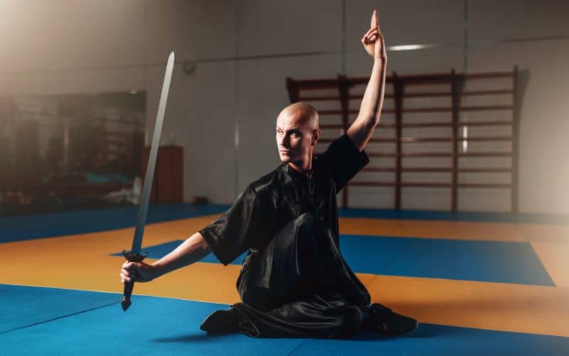 Homme au crâne rasé, en tenue de Taï Chi toute noire, pratiquant le Taï Chi sur un tapis dans une salle de sport, muni d'un sabre.